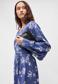 Μπλε φλοράλ φόρεμα με μακριά, φουσκωτά μανίκια, V-ντεκολτέ, ζωνάκι και ροή στη σιλουέτα. Μαύρη τσάντα κρατημένη στα χέρια του μοντέλου.