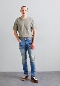 Light gray cotton t-shirt, faded blue denim jeans with a slim fit, and brown sneakers with white and black accents. Standing against a neutral background.
