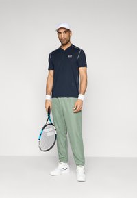 Μπλε ναυτικό πόλο τένις με άσπρες ρίγες, ανοιχτό πράσινο παντελόνι φόρμας και λευκά αθλητικά παπούτσια. Περιλαμβάνει ρακέτα τένις και λευκό καπέλο.