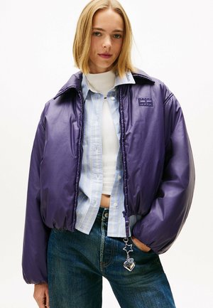 Giacca viola soffice con una texture lucida, colletto grande e orlo elastico, indossata sopra una camicia a righe blu chiaro, con un charm sulla cerniera.