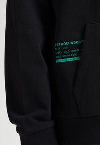 Sudadera negra con bolsillo tipo canguro, puños acanalados y un estampado texturizado con texto en color azul verdoso que detalla la marca y la información de la dirección.