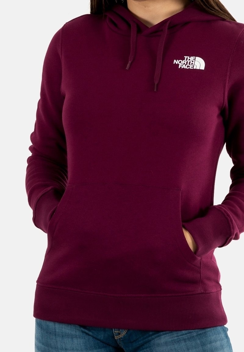 Vrouw draagt een bordeauxrode The North Face hoodie met een voorzak en een capuchon met trekkoord, handen in de zakken, gecombineerd met een blauwe jeans.