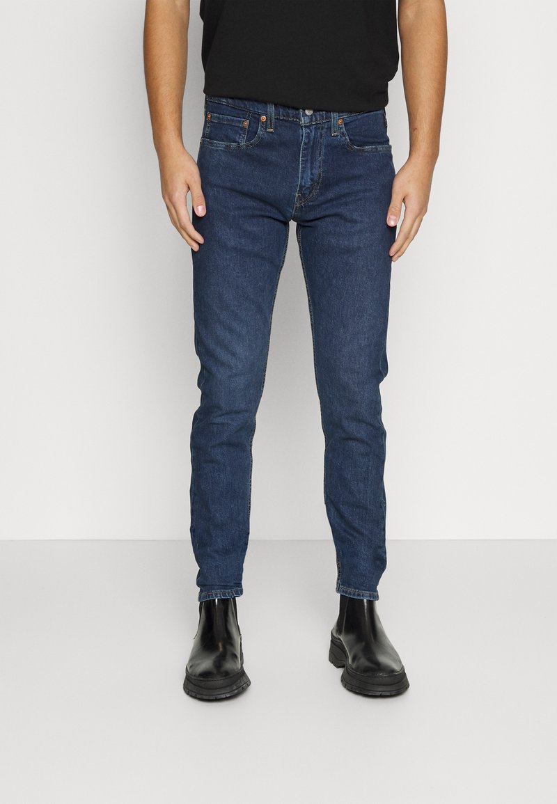 Tummansiniset farkut slim fit -mallilla, joissa on viisi taskua, nappisuljenta ja tikkauskoristeet. Yhdistetty mustiin nilkkureihin.