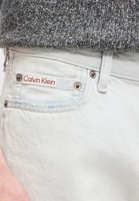 Κλείσιμο τσέπης ανοιχτόχρωμου τζιν με κόκκινη ετικέτα «Calvin Klein» και μέρος γκρι πλεκτού πουλόβερ από πάνω.