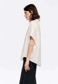 White short-sleeve button-up shirt with a textured fabric, featuring a loose fit and side slits, worn with a black skirt.