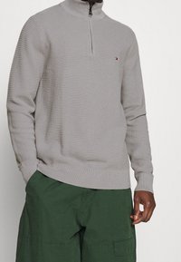 Pullover grigio con zip, dotato di un motivo testurizzato e colletto alto, caratterizzato da un piccolo logo, abbinato a pantaloni cargo verdi.