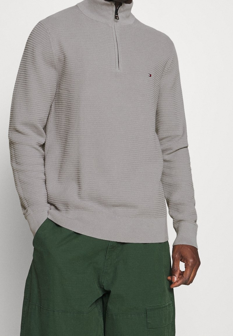 Pullover grigio con zip, dotato di un motivo testurizzato e colletto alto, caratterizzato da un piccolo logo, abbinato a pantaloni cargo verdi.