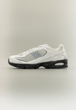 Hvid New Balance sneaker med grå mesh paneler, sort sål og "N" logo på siden, vist mod en ensfarvet baggrund.