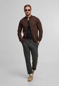 Brauner Strickcardigan mit hohem Kragen, schwarzes, eng anliegendes Hemd, graue Hose mit umgeschlagenen Säumen und beige Schuhe. Die Accessoires umfassen runde Sonnenbrillen.