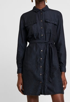 Kobieta nosząca ciemną sukienkę koszulową z denimu z długimi rękawami, zapinaną z przodu na guziki, z kieszeniami na piersi i przewiązanym paskiem w talii.