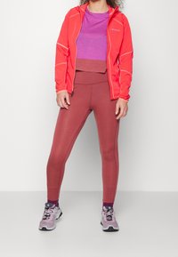 Nainen, jolla on yllään violetti paita, korallinvärinen takki, ruusunkirjavat legginsit ja vaaleanvioletit vaelluskengät, seisoo tasaisella valkoisella taustalla.