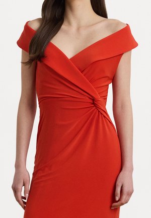 Vrouw draagt een aansluitende rode off-shoulder jurk met een wikkelstijl aan de voorkant en geplooide details in de taille, staand tegen een effen achtergrond.