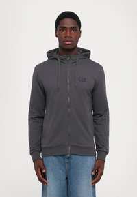 Gray zip-up hoodie with a drawstring hood. Features a small black logo on the chest, ribbed cuffs, and a smooth fabric texture.