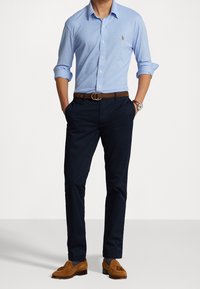 Camicia azzurro chiaro con bottoni, pantaloni blu navy, cintura marrone e mocassini in suede marroni. La camicia presenta un piccolo logo sul petto.