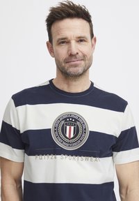 Navy ja valge triibuline T-särk, mis on valmistatud puuvillast, ümmarguse kaelusega ja laurel disainiga insignia ning tekstiga "FQ1924 SPORTSWEAR".