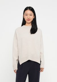 Mlada ženska z dolgimi črnimi lasmi, oblečena v bež oversized pulover in temne hlače, stoji proti enobarvni beli ozadju.