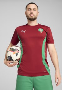 Cămașă de fotbal roșie și verde cu mâneci scurte, având un logo pe piept, asortată cu păshorturi verzi și o minge de fotbal cu model multicolor.