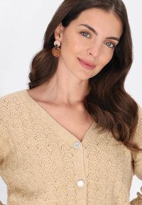 Beżowy sweter dzianinowy z dekoltem w kształcie litery V, z teksturowanym wzorem i srebrnymi guzikami. Kolczyki są okrągłe, z pomarańczowymi i beżowymi akcentami.