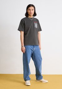 T-shirt gris surdimensionné avec imprimé graphique sur la poitrine, associé à un jean en denim bleu ample et des baskets beiges sur un sol jaune.