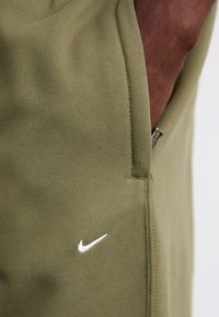 Calças de treino Nike verde oliva com textura canelada, bolsos laterais e um pequeno detalhe do logotipo branco na parte inferior da perna. Detalhe de fecho visível.
