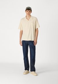 Polo beige de manga corta con un bolsillo en el pecho, combinado con jeans azul oscuro y zapatillas color crema. El modelo lleva una gorra gris claro a juego.
