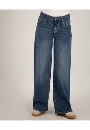 Blauwe denim jeans met wijde pijpen, gedragen met witte sneakers, getoond van taille tot voeten tegen een effen achtergrond.