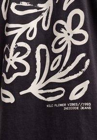 Schwarzer Stoff mit weißen Blumen- und Blattmustern, mit dem Text "WILD FLOWER VIBES/1993 INDICODE JEANS", der unter dem Design gedruckt ist.