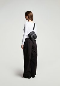 Svart väska med flera fack och justerbar rem som bärs som crossbody. Modell i åtsittande vit långärmad topp och lösa svarta byxor.