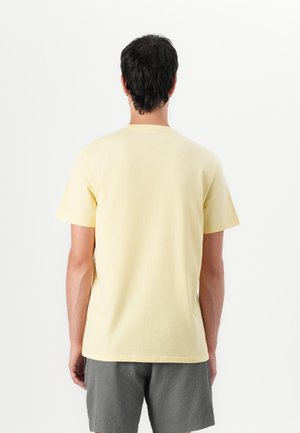 Mand iført en ensfarvet lysegul t-shirt med korte ærmer og grå shorts, stående med ryggen mod kameraet foran en ensfarvet hvid baggrund.