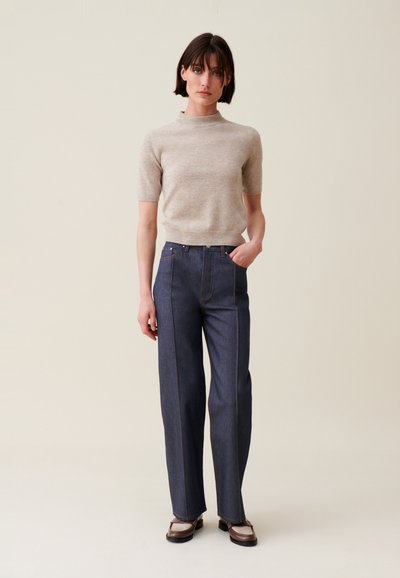 Maglione beige a maniche corte abbinato a pantaloni in denim scuri a vita alta con cuciture visibili e una vestibilità rilassata, completato da scarpe marroni.