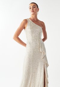 Vestido de un solo hombro con cuentas en beige claro, con un volado lateral, que presenta una abertura alta en el muslo y una silueta ajustada.