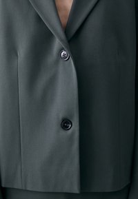 Dark green blazer featuring a structured design, single-breasted front, two black buttons, and smooth fabric texture.