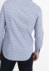 Long-sleeve shirt featuring a blue floral pattern on a white background, with a curved hem and buttoned cuff, made of lightweight fabric.