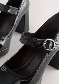 Décolleté Mary Jane in vernice nera con perforazioni stile brogue e cinturino con fibbia argentata su tacchi alti robusti.
