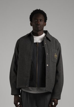 Un homme porte une veste anthracite boutonnée sur le devant, par-dessus une couche noire zippée et un T-shirt blanc, sur un fond gris uni.