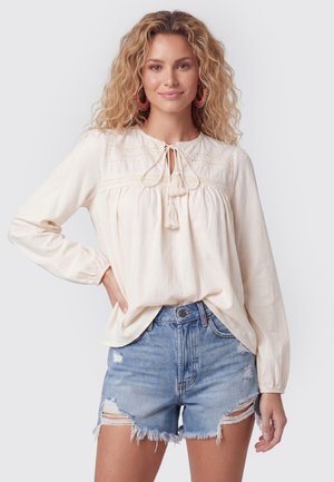 Frau mit lockigem blondem Haar, die eine cremefarbene Langarmbluse mit Fransen und zerrissene hellblaue Jeansshorts trägt, lächelt und posiert mit der Hand in der Hüfte.