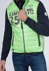 Jasnozielona pikowana kamizelka puchowa z czarnymi akcentami, zamkiem z przodu oraz wyrazistym nadrukiem "25" i "CAMP DAVID" na piersi.
