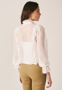 Femme portant une blouse blanche transparente avec un col et des manches à volants, associée à un pantalon moutarde orné de motifs floraux brodés.