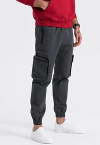 Dunkelgraue Cargo-Hosen mit elastischen Bündchen, ausgestattet mit Seitentaschen und einer Reißverschlusstasche. Getragen mit einem roten Sweatshirt und weißen Sneakers.