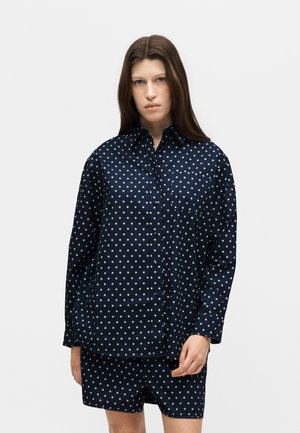 Mladá žena s dlhými hnedými vlasmi nosí námornícky modrú bodkovanú košeľu na gombíky a zodpovedajúce šortky, stojí na jednoduchom svetlom pozadí.