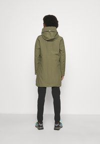Olive green waterproof parka with a hood, cinched waist, and two side pockets. Worn over black pants and gray athletic shoes.