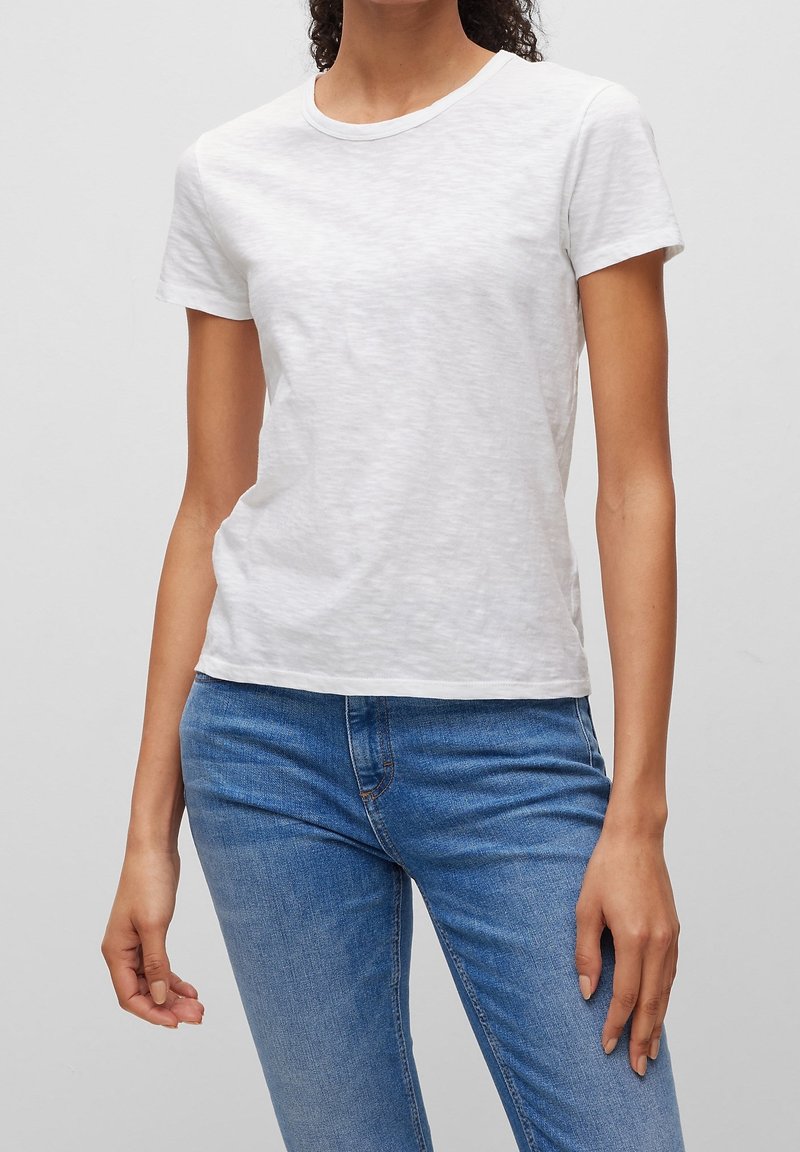 Femme portant un t-shirt blanc uni à manches courtes et un jean denim bleu, debout devant un fond gris clair uni.