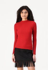 Rode geribbelde langemouwen top met een geschulpte halslijn, gecombineerd met een zwarte fringed rok, die textuur en ontwerpaccentueert.