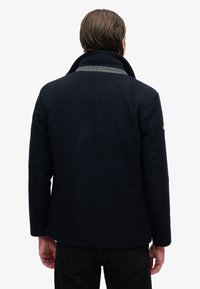 Chaqueta de lana azul marino con cuello alto, forro interior gris y hombros estructurados. Presenta un ajuste entallado y un diseño minimalista.