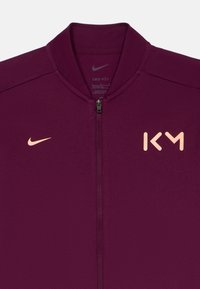 Μάυρη ζακέτα Nike με ψηλό γιακά, μπροστινό φερμουάρ και αντίθετο φωτεινό πορτοκαλί λογότυπο "KM" στην αριστερή πλευρά και το λογότυπο Nike στον θώρακα.