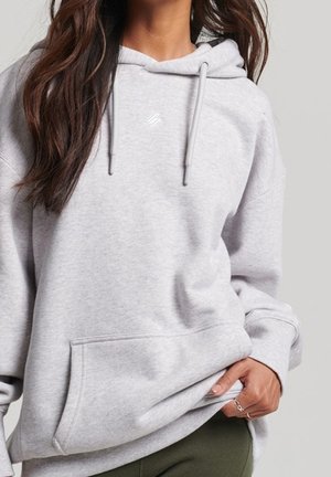 Vrouw draagt lichtgrijze capuchontrui met voorzak, losse pasvorm en subtiel logo op de borst, hand trekt zoom iets omhoog.