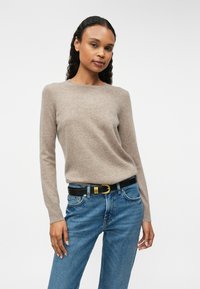 Pull en tricot marron clair avec un col rond, associé à un jean bleu et une ceinture noire avec des éléments en or. Texture lisse, design ajusté.