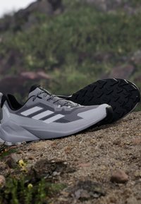 Zapatillas de correr grises con parte superior texturizada, suela de caucho negra y acentos de rayas blancas. Presentan cierre de cordones y talón reforzado.