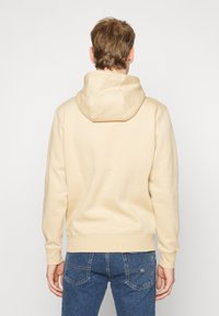 Sudadera con capucha beige con bolsillo tipo canguro al frente, puños y dobladillo acanalados. Textura suave y capucha ajustable con cordón. Pantalones vaqueros azules puestos.