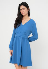 Μπλε φόρεμα με μακριά μανίκια, V-λαιμόκοψη, κουμπιά στο μπροστινό μέρος και ζώνη στη μέση. Το ύφασμα έχει υφή και η φούστα είναι φαρδιά.